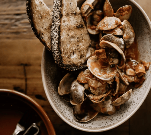 A bowl of clams in a savory sauce, served with toasted bread on the side, on a wooden table.
