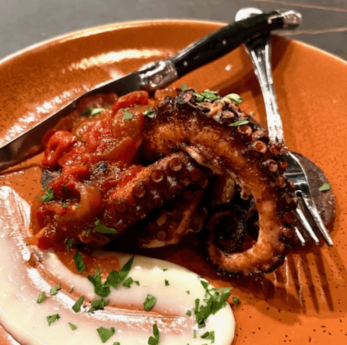 Grilled octopus on a plate with tomato sauce, garnished with herbs, alongside a creamy sauce and cutlery.