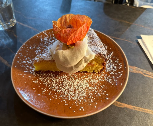 A slice of cake topped with whipped cream and a flower, dusted with powdered sugar on a brown plate.