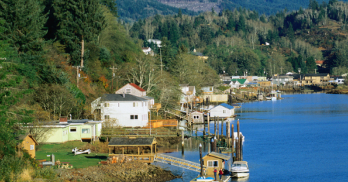 Scenic view of a coastal town with houses along a river, surrounded by mountains and lush greenery.