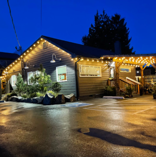 A cozy building illuminated with string lights at dusk, surrounded by greenery and a wet parking lot.