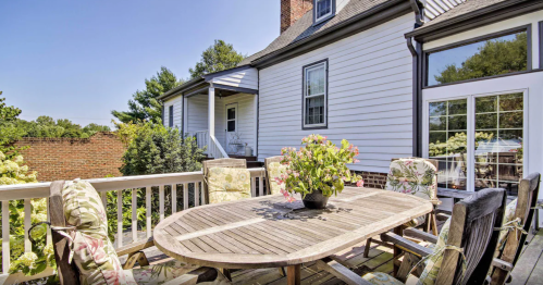 A wooden dining table with floral chairs on a sunny deck, surrounded by greenery and a view of a house.