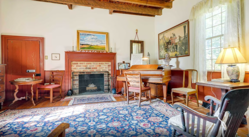 Cozy room with a fireplace, wooden furniture, a piano, and a patterned rug, illuminated by natural light.