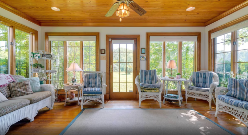 Bright sunroom with wicker furniture, large windows, and a wooden ceiling, overlooking a green outdoor view.