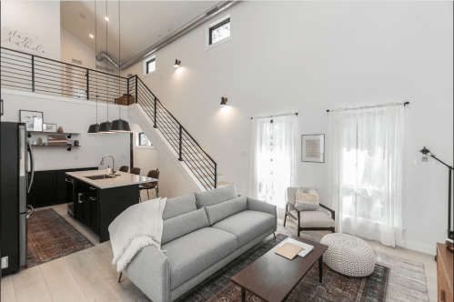 Modern living room with a gray sofa, staircase, and kitchen area, featuring large windows and bright, airy decor.