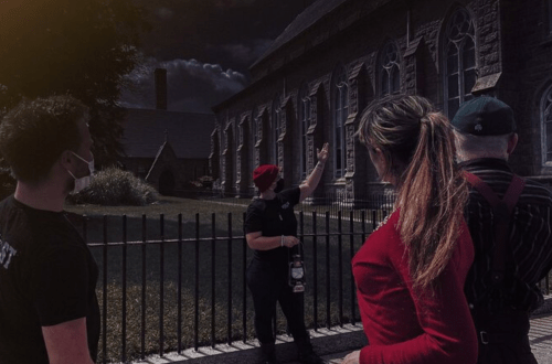 A group of people listens to a guide pointing at a historic building under a dark sky.