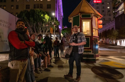 A group of people gathered on a city street at night, with a colorful kiosk and buildings illuminated in the background.