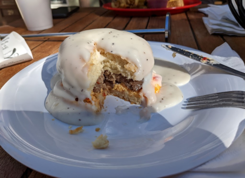 A partially eaten biscuit sandwich with sausage and creamy gravy on a white plate, set on a wooden table.