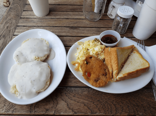 A plate of biscuits with gravy, scrambled eggs, a fried patty, toast, and a small dish of jelly on a wooden table.