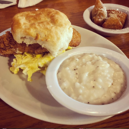 A plate with a biscuit sandwich filled with fried chicken and scrambled eggs, alongside a bowl of gravy and a dish of pastries.