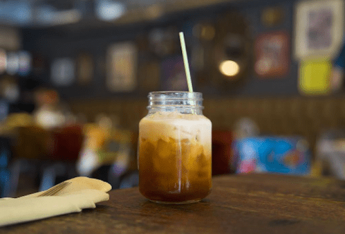 A jar of iced coffee with a straw sits on a wooden table in a cozy café setting.