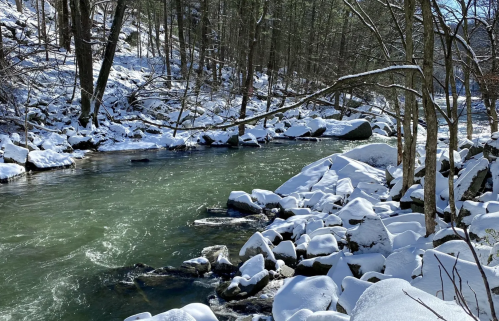 A serene winter scene featuring a flowing river surrounded by snow-covered rocks and trees.