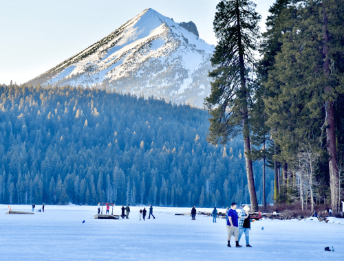 People walking on a frozen lake with a snow-capped mountain and tall trees in the background.