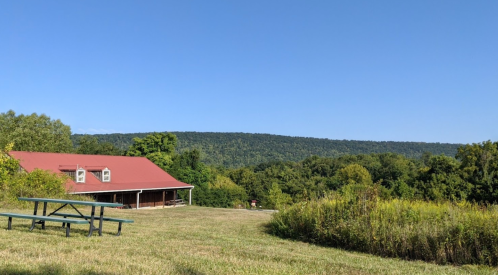 A red-roofed building sits in a grassy area, surrounded by trees and rolling hills under a clear blue sky.