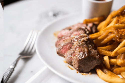 A plate featuring sliced steak with a side of golden French fries and a small cup of dipping sauce.