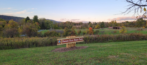 Sign for Sweet Run State Park in a scenic landscape with trees and rolling hills under a colorful sky.