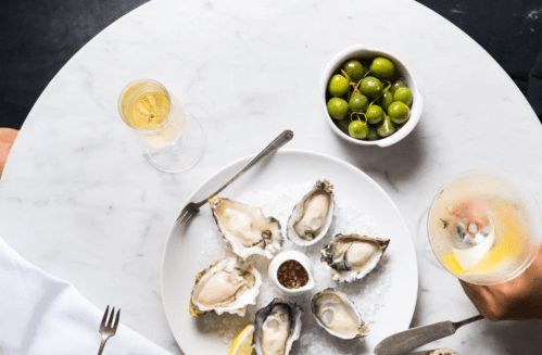A marble table with a plate of oysters on ice, a bowl of green olives, and two glasses of sparkling drinks.
