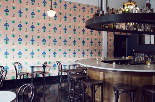 A stylish bar with a curved counter, surrounded by wooden stools and floral wallpaper in soft pastel colors.