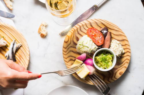 A wooden plate with assorted appetizers, including vegetables, cheese, and a small bowl of dip, beside a glass of wine.