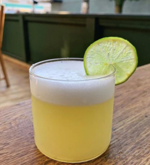 A refreshing yellow cocktail with a frothy top, garnished with a lime slice, served in a clear glass on a wooden table.