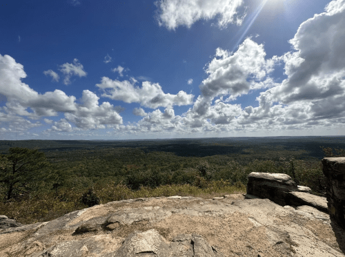 A panoramic view from a rocky overlook, showcasing a lush green landscape under a bright, cloudy sky.