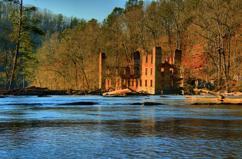 Ruins of a brick building by a calm river, surrounded by trees and autumn foliage.