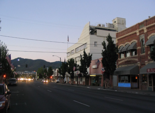 Quiet street scene at dusk, featuring buildings, trees, and mountains in the background, with streetlights and flags.