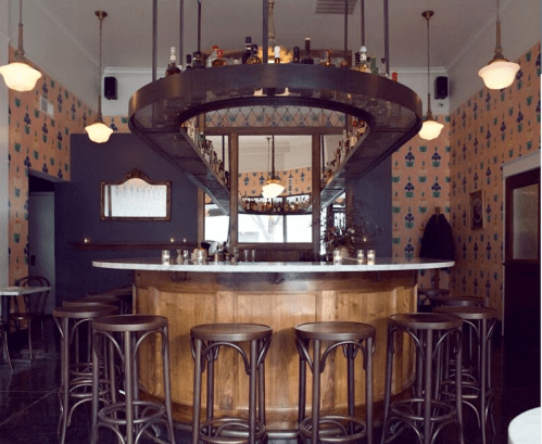 A stylish bar with a curved wooden counter, high stools, and patterned wallpaper in a cozy setting.