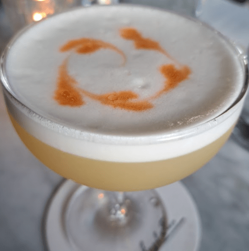 A frothy cocktail in a coupe glass, featuring a swirl of orange garnish on top of a pale yellow drink.