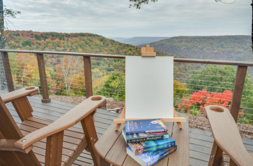 A wooden deck with two chairs, an empty easel, and a stack of books, overlooking a colorful autumn landscape.