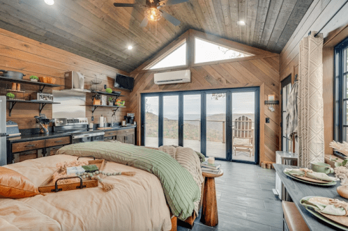 Cozy cabin interior with a bed, kitchen area, and large windows showcasing a scenic view. Warm wood accents throughout.