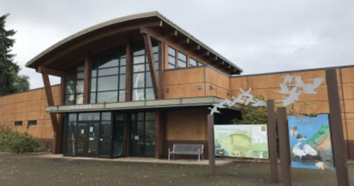A modern building with a wooden roof and large windows, featuring a sign and artwork outside.