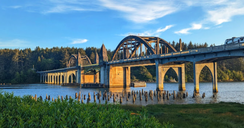 A bridge spans a river, surrounded by trees and grassy banks, under a blue sky with wispy clouds.