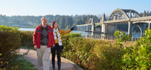 A couple walks along a path by a river, pointing at a bridge in the background surrounded by trees and fog.