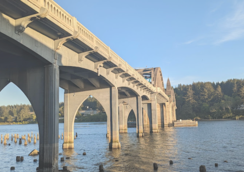 A concrete bridge spans over a calm river, with trees lining the shore under a clear blue sky.