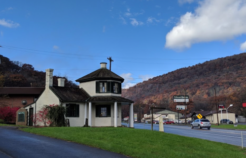 A quaint two-story building with a round turret, set against a backdrop of hills and a clear blue sky.