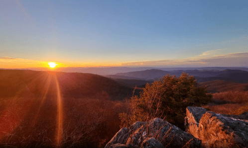 A stunning sunset over rolling mountains, with warm hues illuminating the sky and rocky foreground.
