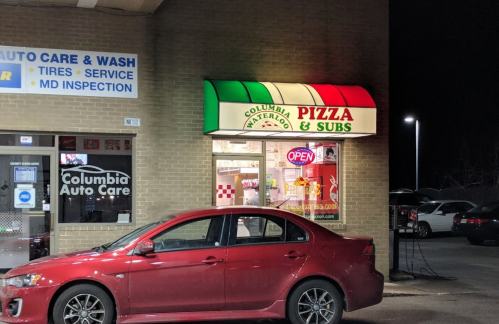 A pizza and subs shop with a colorful sign, next to an auto care service, illuminated at night. A red car is parked outside.