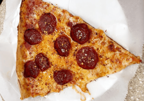 A slice of pepperoni pizza with melted cheese and spices on a white paper plate.