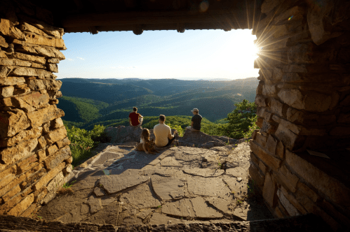 Four people and a dog sit on a rocky ledge, overlooking a scenic valley at sunset.