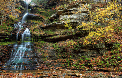 A serene waterfall cascading down rocky cliffs, surrounded by autumn foliage and a tranquil pool below.