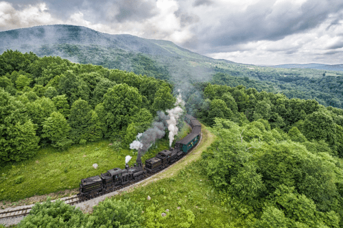 A steam train winds through lush green hills under a cloudy sky, releasing plumes of smoke.