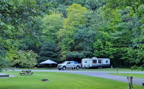 A blue truck towing a camper parked near a picnic area, surrounded by lush green trees.
