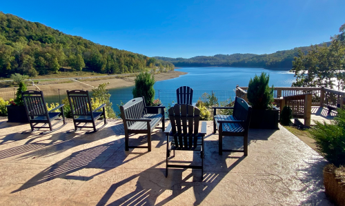 A serene lakeside view with empty chairs facing the water, surrounded by trees and a clear blue sky.