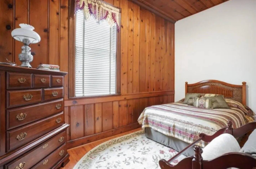 Cozy bedroom with wooden walls, a bed with striped bedding, a dresser, and a decorative rug. Natural light from a window.