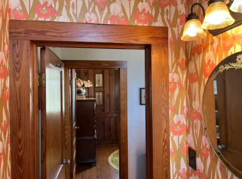 A hallway with floral wallpaper, wooden trim, and a round mirror, leading to a room with wooden furniture.