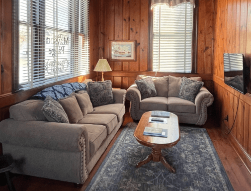 Cozy living room with wooden walls, two sofas, a coffee table, and a lamp, featuring natural light from large windows.