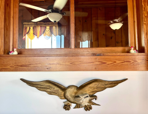 A wooden eagle sculpture hangs on a wall beneath a window with decorative curtains and two small figurines.