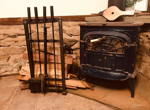 A vintage wood stove beside a stack of firewood and fireplace tools against a stone wall.