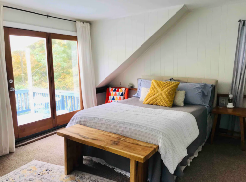 Cozy bedroom with a sloped ceiling, a bed with decorative pillows, and a wooden bench by a sliding glass door.